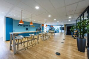 Moderne bedrijfskeuken en ontmoetingsruimte bij Ayvens aan de Johan Huizingalaan, met hoge tafels, barkrukken en oranje hanglampen.